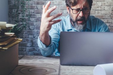  Video görüşmesinde iş adamı dizüstü bilgisayara kızgın bağırıyor. Bir yetişkin erkek, internet üzerinden iş yapıyor ve sorunları yönetiyor. Bilgisayarın önünde yorgun düşmüş bir işçi kızgın bir şekilde bağırıyor.