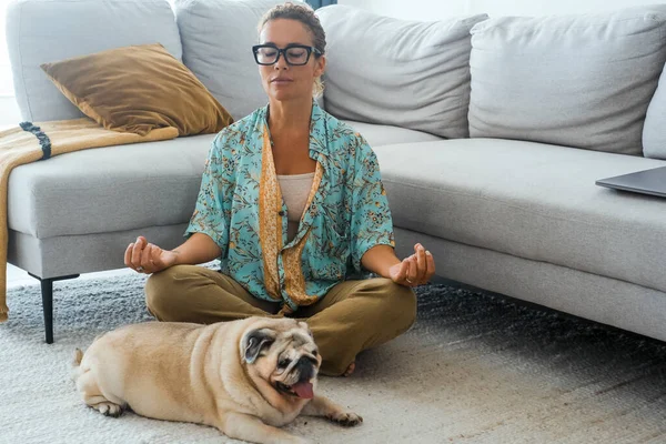 woman having relax at home with yoga routine lotus position in living room with her best friend dog laying near her. Concept of easy and healthy mental lifestyle people. Dog owner. Wellbeing lady