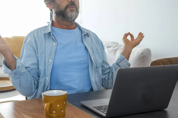 Concept of meditation and relaxation during online work with computer. Stress and problems on business job. One man doing yoga lotus position and breathe in front of an open laptop on the table at home