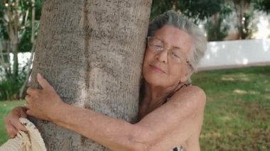  90 'ların yaşlı kadını bir ağaca sevgiyle sarılıyor. Parktaki yaşlı bayanın mutlu ve huzurlu ifadesi. Sağlıklı emekliler yaşam tarzı kavramı