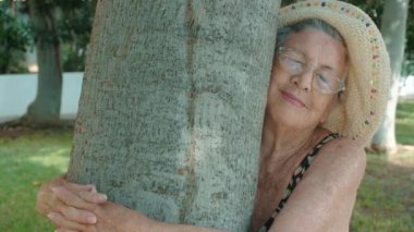  90 'ların yaşlı kadını bir ağaca sevgiyle sarılıyor. Parktaki yaşlı bayanın mutlu ve huzurlu ifadesi. Sağlıklı emekliler yaşam tarzı kavramı