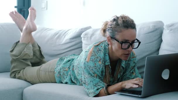 屋内レジャー活動の女性そしてコンピュータ無線関係 若い成人女性の1人は リビングルームの自宅のソファーに彼女の胃の上に横たわるノートパソコンを使用しています メガネを着て執筆する女性 — ストック動画