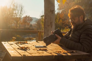 Adam, sonbahar sezonunda, açık havada tahta bir masanın üzerinde büyük bir dslr kamera kullanıyor. Parkta sarı yapraklar. Fotoğrafçı refleksteki resimleri kontrol ediyor. Açık hava aktivitesi yapan insanlar. Modern çalışma
