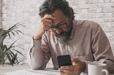 Ofisteki adam endişeli bir ifadeyle cep telefonu kullanıyor ve düşünceleri ön tarafına dokunuyor. Modern iş hayatındaki iş ve sorunlar. Masada akıllı telefon kullanmak. Küçük işletme yetişkin