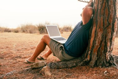 Dışarıda bilgisayar ve kablosuz bağlantıyla çalışan insanlar. Sırtını dizüstü bilgisayarı bacaklarında bir ağaca dayayan bir adam. Bitiş projesi ve dijital göçebe yaşam tarzı kavramı. Modern iş