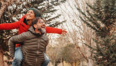 Happy adult couple of tourist have fun and enjoy outdoor leisure activity playing and laughing a lot.  man carrying woman in piggyback on his back. Female outstretching arms celebrating nature love clipart