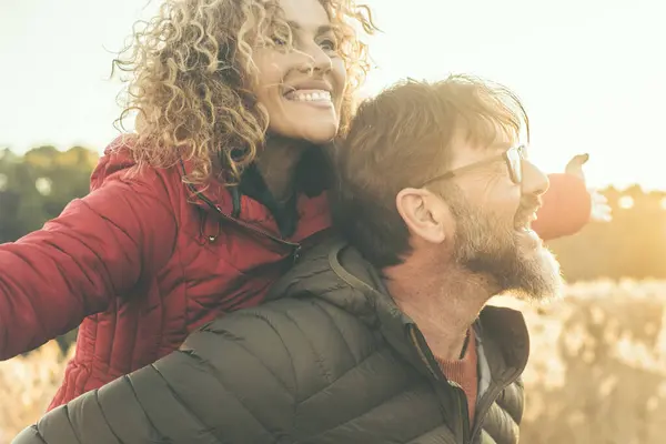 Happy couple have fun together in outdoor leisure activity in nature field during sunset time and golden hours light. Man carry woman on his back. People enjoying life and laughing. Winter autumn day