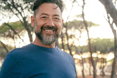 Gülümseyen ve kameraya bakan mutlu mutlu adam portresi. Arka plandaki ağaçlar doğa parkı. İnsanlar dışarıda boş vakit geçirmekten hoşlanıyorlar. Yetişkin bir erkek sana bakıyor. Boşluğu kopyala