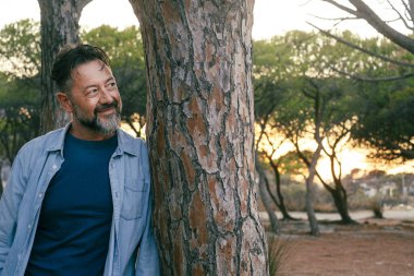 Adam parkta bir ağaca yaslanmış, yüzünde gülümseme ve dingin bir ifadeyle yan tarafa bakıyor. İnsanlar hafta sonu aktivitelerini ve boş zamanlarını severler. Sıradan kot elbiseler. Modern son sınıf öğrencisi erkek