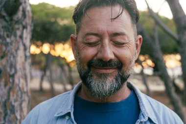 Kameraya gülümseyen sakallı ve kapalı gözlü olgun bir adamın portresi. Refah ve ruh sağlığı konsepti mutluluk yaşam tarzı. İç yaşam dengesi pozitif. Açık havada gezen yetişkin erkekler