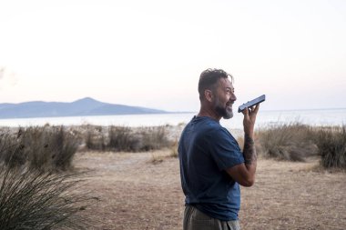 Turist açık hava seyahatlerinde tek başına boş zaman aktivitelerinde konuşuyor. Arka planda okyanus ve sahil adası var. Maceracı hayat tarzı. Dışarıda dolaşıyordum. Hücre hoparlörü tutuluyor