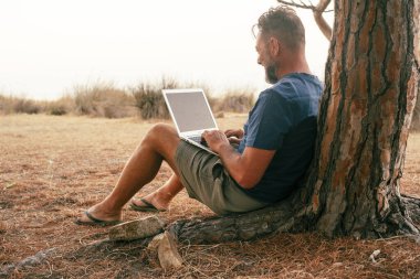 Modern gezgin serbest işçi yaşam tarzı dizüstü bilgisayarda çalışıyor bir ağaca karşı oturuyor ve doğanın tadını çıkarıyor. Bilgisayar bağlantısı ve küçük çevrimiçi iş teknolojisi dışında.