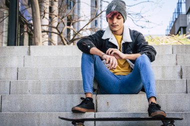 Şehirdeki parkta tek başına oturan genç bir çocuk aşağıya bakıyor. Kaykay nakliyesi. Gençlerin yaşam tarzı. Şehir hayatı insanları. Sıradan kot pantolon, alternatif saç stili. Açık havada 18 'lik.