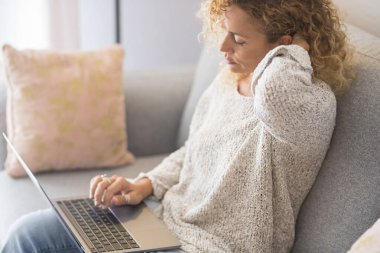  Kadın evinde kanepede oturuyor. Dizüstü bilgisayar kullanıyor. Yorgun ve rahat. Kablosuz bilgisayar insanları çalıştırır. Yetişkin bir kadın internette tek başına sörf yapıyor. Ağrı ve kötü duruş için boyuna dokunmak sağlıksız.