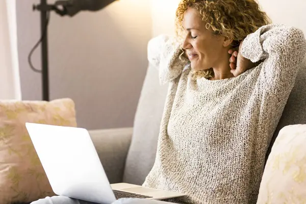 woman finish to use laptop at home sitting on the sofa stretching neck and shoulder back with a satisfied smile. Portrait of modern adult female people using computer sitting on the sofa. Indoor.