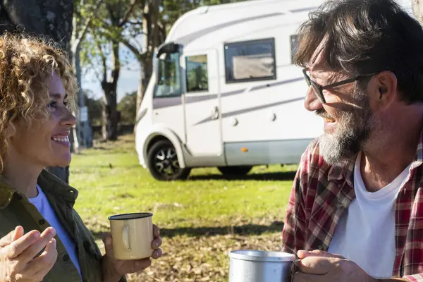 Happy couple of traveler tourist enjoy relax and drinking coffee during travel pause in outdoor at the park with modern rv motorhome vehicle in background under the trees. Autumn journey adventure
