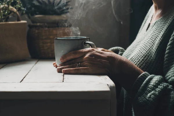 Close up of woman hands holding warm beverage cup at home indoor with smoke and dark light. Relaxation leisure activity. Drinking healthy beverage. Coffee time. Green mood color. Serene lifestyle lady