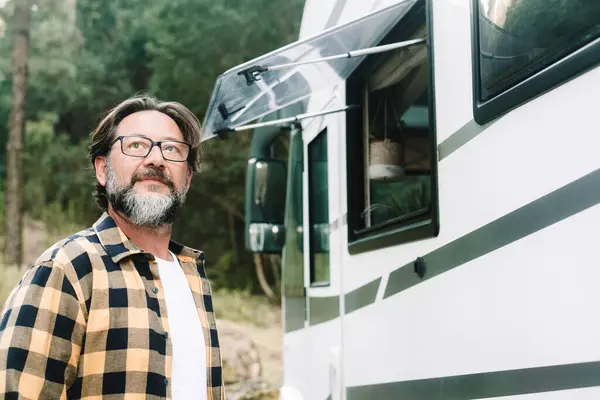 man outside a camper van motorhome rv vehicle looking the outdoors in mountain forest woods scenic destination. Alternative travel and lifestyle people. Mature male with beard and camper van