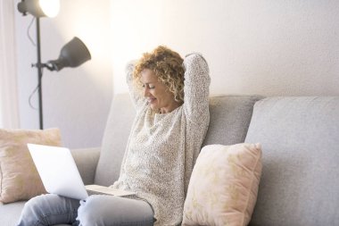 Aydınlık evdeki koltukta dizüstü bilgisayar kullanan bir kadın. İnsanlar ve teknoloji çevrimiçi. Modern kadın bilgisayarı izliyor, sırtını ve omuzlarını dinlendiriyor ve kollarını başının altından kaldırıyor.