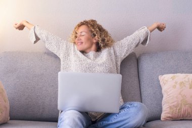  Kanepede oturup dizüstü bilgisayar kullanmak için kollarını uzatan mutlu bir kadın. Çevrimiçi eğlence aktivitesi kavramı. Yorgun kadın gülümsüyor ve başarı yaşam tarzının tadını çıkarıyor. Modern insanlar çevrimiçi