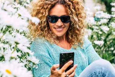 Açık havada cep telefonu kullanan beyaz, sevimli bir kadın. Etrafta papatyalarla oturuyor. Mutlu kadınlar sohbet eder ve boş zamanlarında hücresel cihaza mesaj gönderir.