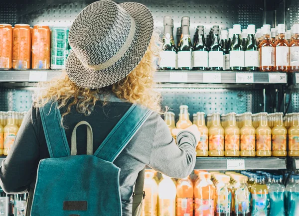 Back view of woman tourist with backpack in front of a window store full of drinks choosing one to buy and drink. Concept of travel solo female people in front of an automatic machine