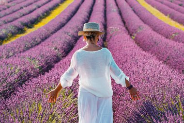 Beyaz elbiseli ve şapkalı bir kadın lavanta menekşe çiçekleriyle yürüyor. Dişi, manzaralı yolculukların tadını çıkarıyor. Gezginler için açık hava aktivitesi. Güzel doğa yazlık bir yer. Zarafet