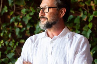 Kolları çapraz ve arka planda yeşil yapraklarla gülümseyen gözlüklü olgun sakallı bir adamın şirket portresi. Kopya alanı olan kendine güvenen yetişkin erkekler. Beyaz gömlekli adam.