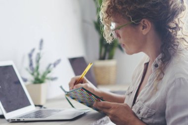 Modern ev hanımı aile ekonomisini evde yönetiyor - bekar bayan not almak ve çalışmak için dizüstü bilgisayar ve kitap kullanıyor - masaüstünde oturup çalışan yetişkin bir kadın.