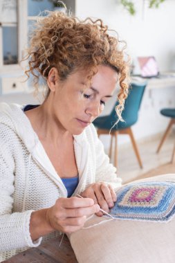 Adult caucasian woman at home doing crochet hobby activity at home decorating a shabby chic white pillow. Living room interior in background. Female people creating woolen decorations