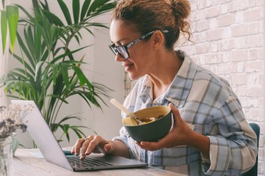 Ofis masasında çalışan bir kadın kasede sağlıklı yiyecekler yiyor. Bağımsız kadın yaşam tarzı, dizüstü bilgisayarları ve online fırsatları kullanarak alternatif iş hayatı. Modern işçi işleri
