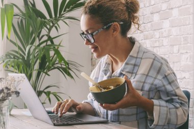 Ofis masasında çalışan bir kadın kasede sağlıklı yiyecekler yiyor. Bağımsız kadın yaşam tarzı, dizüstü bilgisayarları ve online fırsatları kullanarak alternatif iş hayatı. Modern işçi işleri