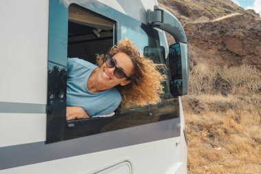 Mutlu ve özgür bekar bir kadın. Kamp arabasının penceresinin önündeki varış yerinin keyfini çıkarıyor ve gülümsüyor. Tatilde seyahat etmek için karavan kiralama fikri. Minibüs hayatı ve şebeke dışı