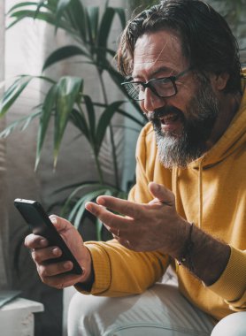 Evde video bağlantısı yapan bir adam telefon kullanarak boş zaman aktivitesi yapıyor. Laptop arka planda. Ev içi aktivitelerdeki insanlar teknoloji ve internet bağlantısı kullanıyorlar. Evde cep telefonundan hoşlanan bir erkek