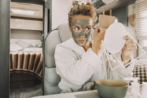 Caucasian woman making beauty skin treatment using cream and mirror sitting and relaxing inside a camper van in travel van life lifestyle. Adult female people have care and use mask for aging