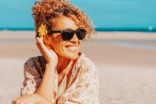 Portrait of happy tourist sitting and smiling at the beach with blue sky and ocean in background. Travel and tourism in summer holiday vacation. Female people with sunglasses on the sand
