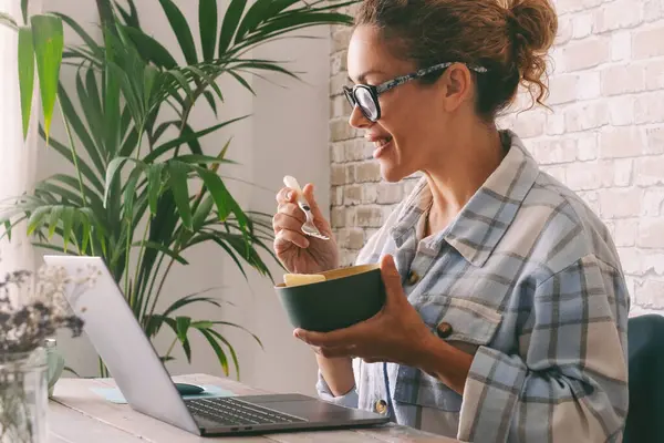 Side view of adult  woman smiling and eating in from of a laptop at the desk at home. Alternative office lifestyle, female people working in video call conference during lunchtime. Modern job