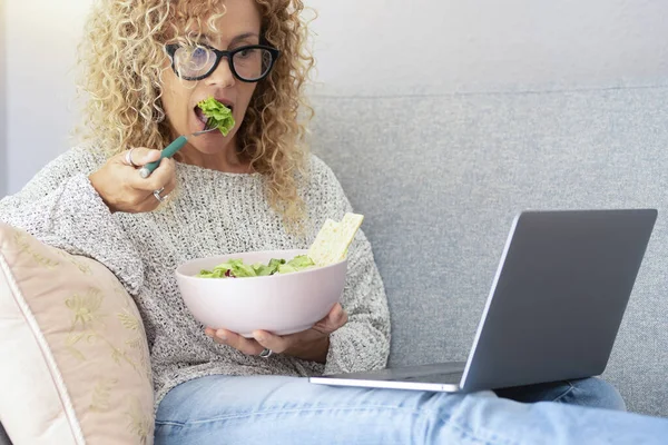 Sağlıklı yaşam tarzı ve modern online faaliyet konsepti. Kanepede oturmuş salata yiyen ve dizüstü bilgisayara bakan bir kadın. Evde tek başına bilgisayar kullanan bir kadın. Kapalı alanda boş zaman web etkinliği