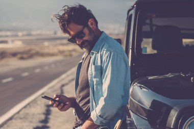Gezgin adam, arka planda uzun yolu olan siyah bir off-road aracına karşı durarak mobil telefon bağlantısı kullanıyor. Maceraperest erkekler için yolculuk ve seyahat yaşam tarzı. Dijital harita yol gezisi kullanımı