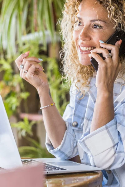 Adult woman calling at the phone sitting on the table. Pretty female people speaking and using mobile cellular and laptop at the desk. Businesswoman manage online ecommerce store for business activity