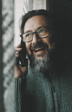 Mutlu, olgun bir genç adam telefonda gülümsüyor ve pencerenin yanında eğleniyor. İnsanlar ve cep telefonu iletişimi kavramı. Erkek kişi arkadaşlarıyla iletişim kurmak için cep telefonu kullanıyor.