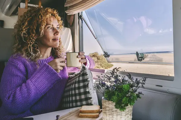 woman alone inside camper van motor home admiring outdoors scenic parking and drinking tea in relax leisure indoor activity. Travel people for vacation and lifestyle van life off grid. Phone table