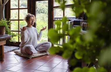 Uzun boylu, beyaz bir kadın mudra jesti yapıyor, lotus pozisyonunda oturuyor evdeki yumuşak minderde oturuyor. Huzurlu kadın derin meditasyon yapıyor, yoga egzersizleri yapıyor..