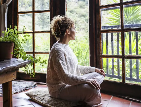 beautiful calm caucasian woman sitting on a comfortable pillow placed on floor at home and feeling grateful and grateful. Happy woman shows gratitude, love and care, looking out the window. concept of faith and relaxation.