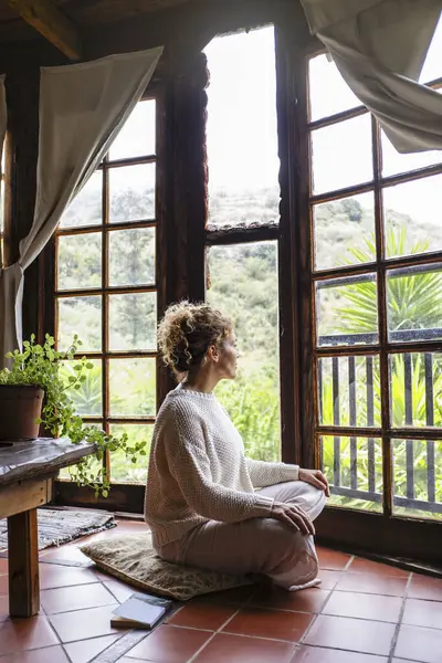 beautiful calm caucasian woman sitting on a comfortable pillow placed on floor at home and feeling grateful and grateful. Happy woman shows gratitude, love and care, looking out the window. concept of faith and relaxation.