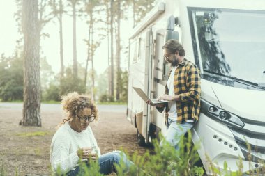Karavan karavanı karavan konsept yaşam tarzında yaşayan insanlar. Bitkilerle rahatlayan bir kadın ve dışarıda dizüstü bilgisayarda çalışan bir adam. Arka planda manzaralı orman ormanı. Gezgin hayatı