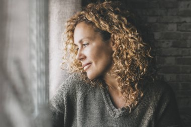 Yetişkin bir kadın, yüzünde barışçıl bir ifade ve küçük bir gülümsemeyle pencereden dışarı bakıyor. Kapalı alandaki kendine güvenen kadınlar boş zamanlarını rahat geçirirler. Hayal kuran ve düşünen bayan
