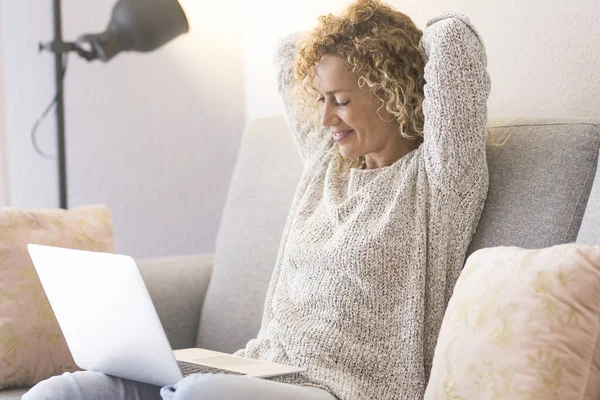一个女人坐在明亮的家沙发上 用笔记本电脑 人和技术在线 现代女性观察电脑 放松后背和肩膀 把胳膊伸到头顶下 — 图库照片