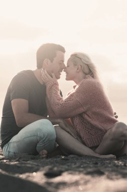 Romantic evening with girl and boy together sitting at the beach on the sand kissing in tenderness. Two people male and female in relationship sitting on the sand and enjoying sunset. Man woman kiss