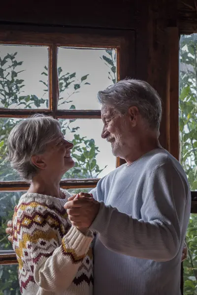 Old man and woman happy couple dancing at home in living room with outdoors background and windows. Senior people enjoy chalet for winter vacation together. Love and life forever retired leisure